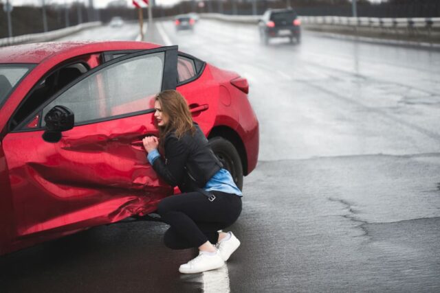 A woman sits beside a damaged car following an accident, reaching out for assistance. Concept of car insurance and seeking help after a collision.