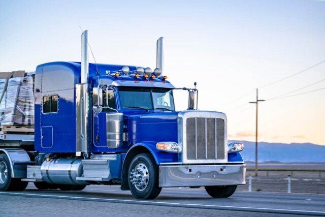 A blue industrial big rig semi-truck with chrome parts hauling secured commercial cargo on a flatbed trailer, driving along a highway in Fort Smith.