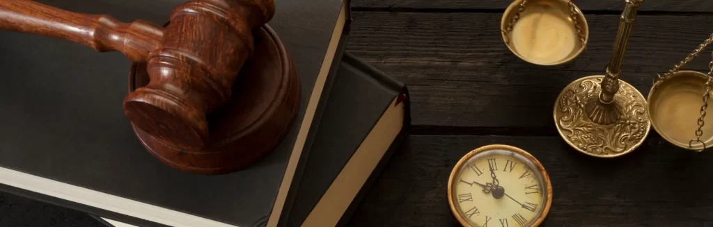A gavel on a book, a vintage clock, and brass scales of justice on a dark wooden table.