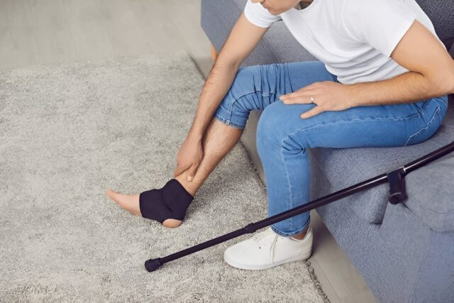 Young man with crutches in casual clothes massaging his injured leg sitting on sofa at home in black bandage.